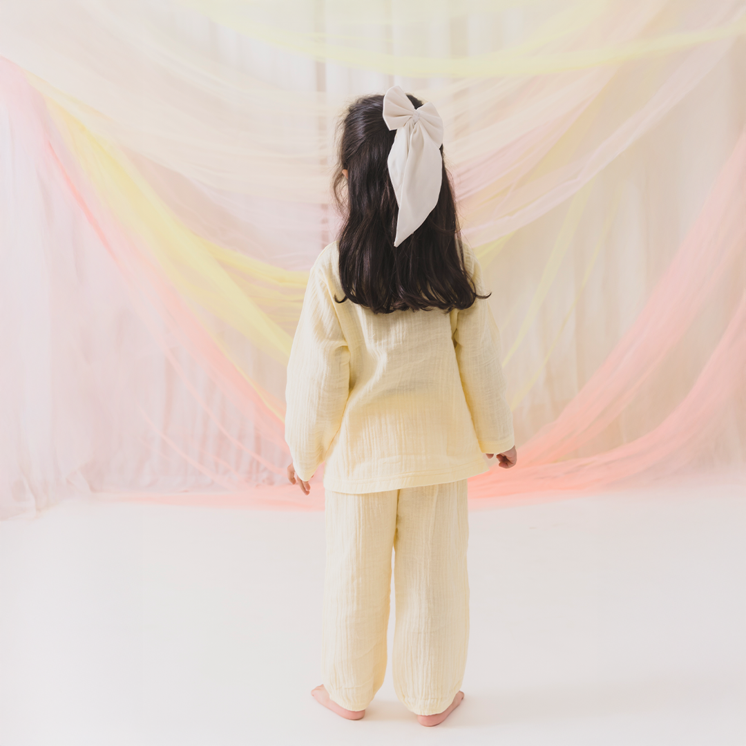 Child wearing a light-colored outfit with a large bow in a softly lit room.