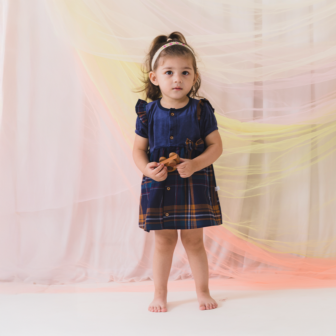 Young girl in a blue dress with plaid skirt standing in front of colorful fabric