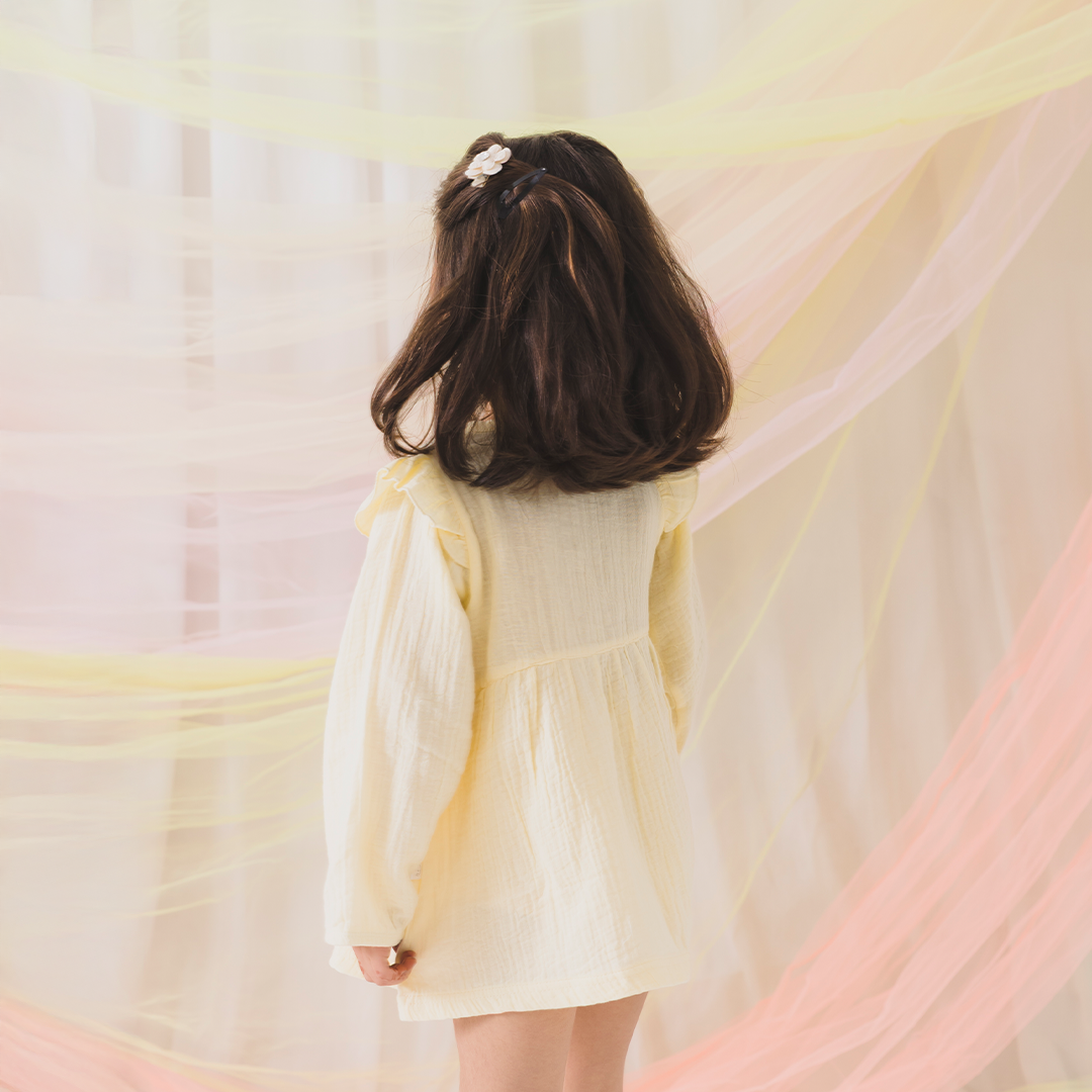 Child in a light yellow dress standing in front of a soft, pastel-colored curtain.