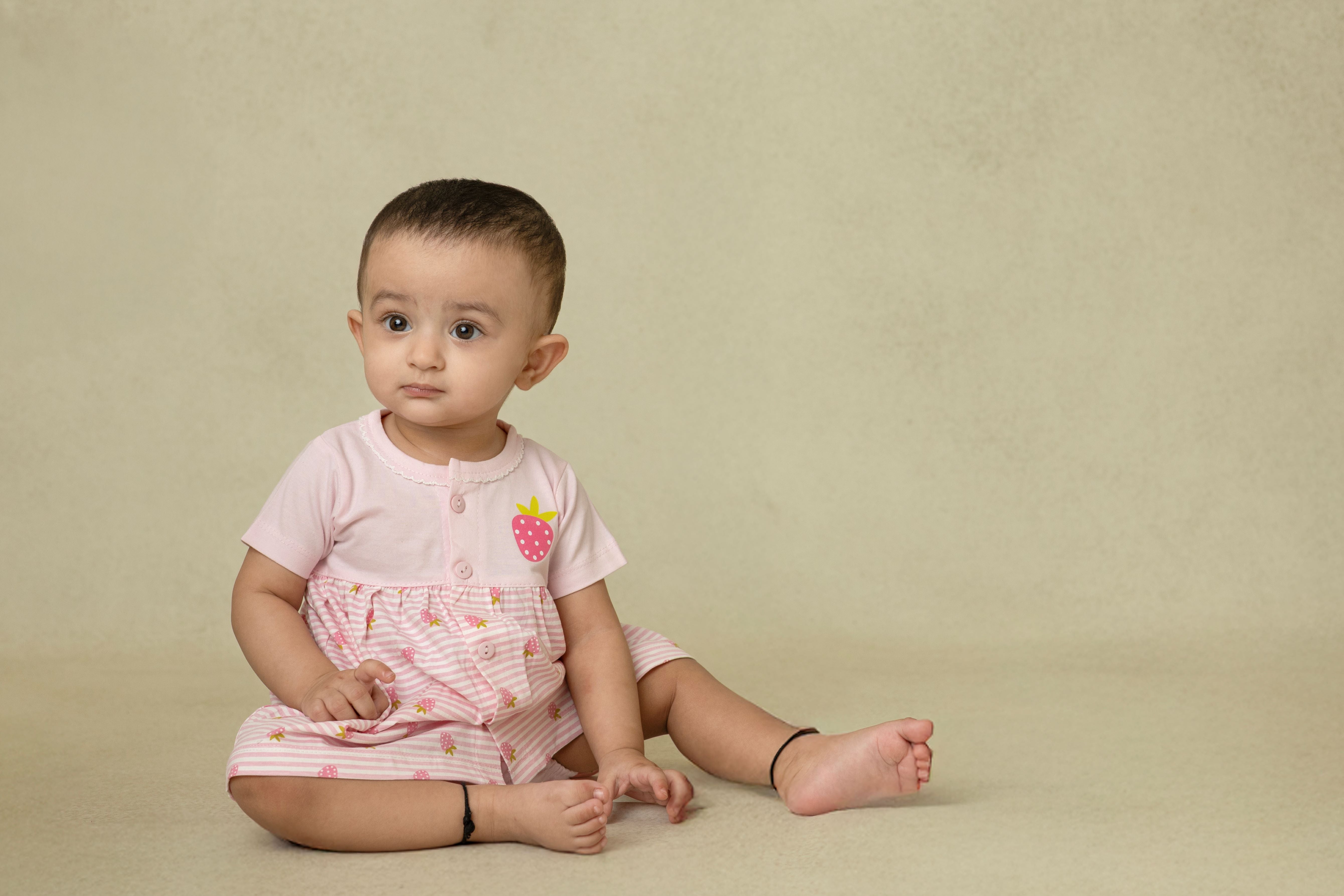 A baby pink dress with strawberry prints, featuring a flowy silhouette and an easy button-down front, worn by a sitting baby.