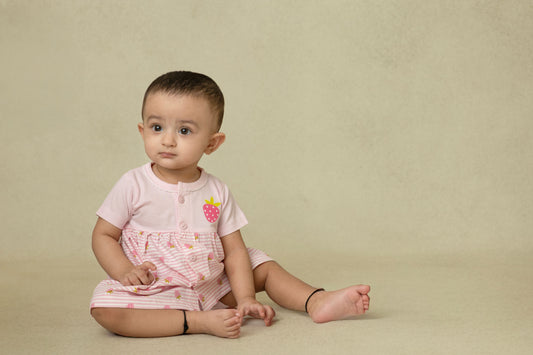 A baby pink dress with strawberry prints, featuring a flowy silhouette and an easy button-down front, worn by a sitting baby.