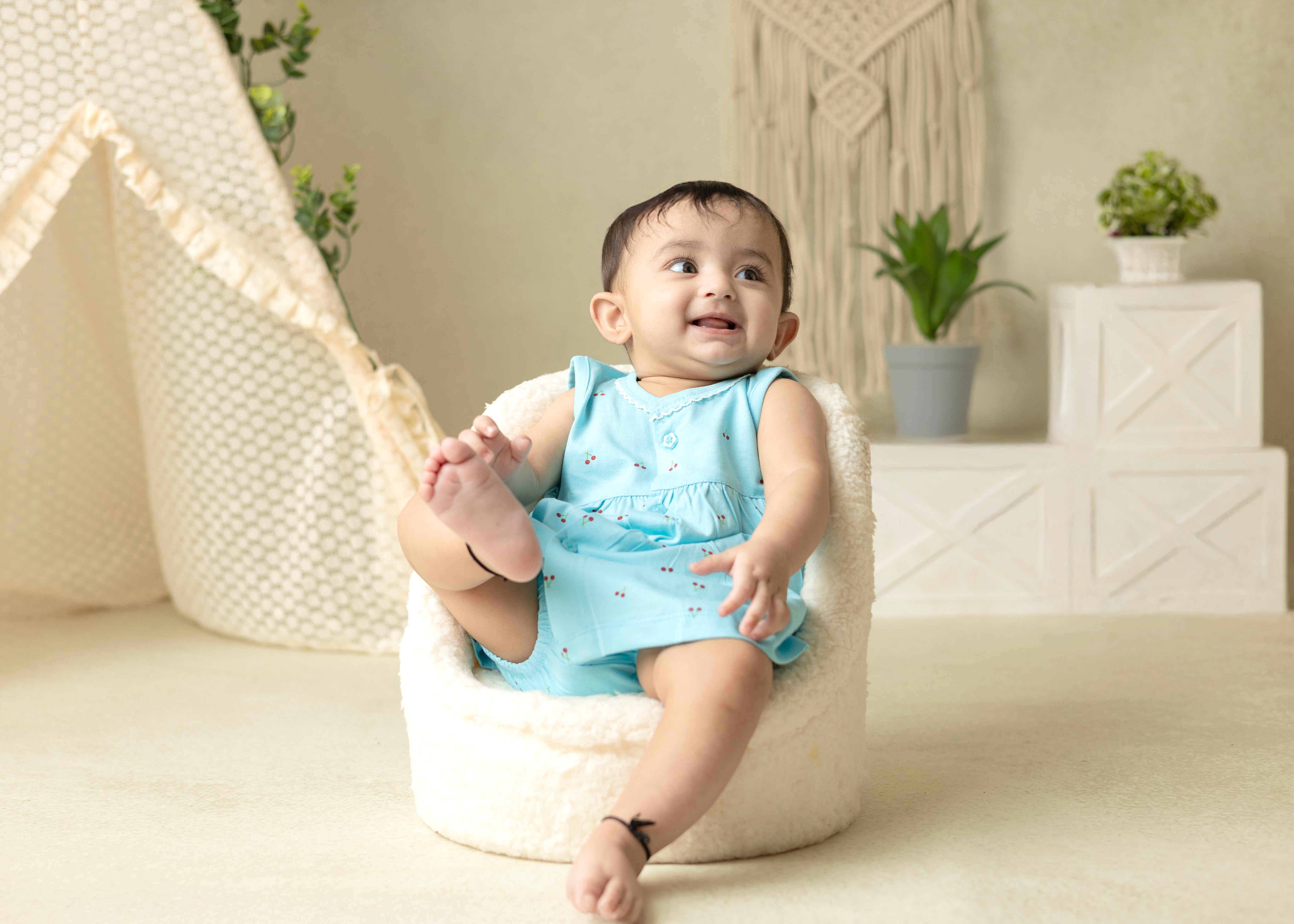A baby wearing a blue sleeveless cotton frock with cherry blossom prints and matching bloomer, sitting in a white bean bag chair.