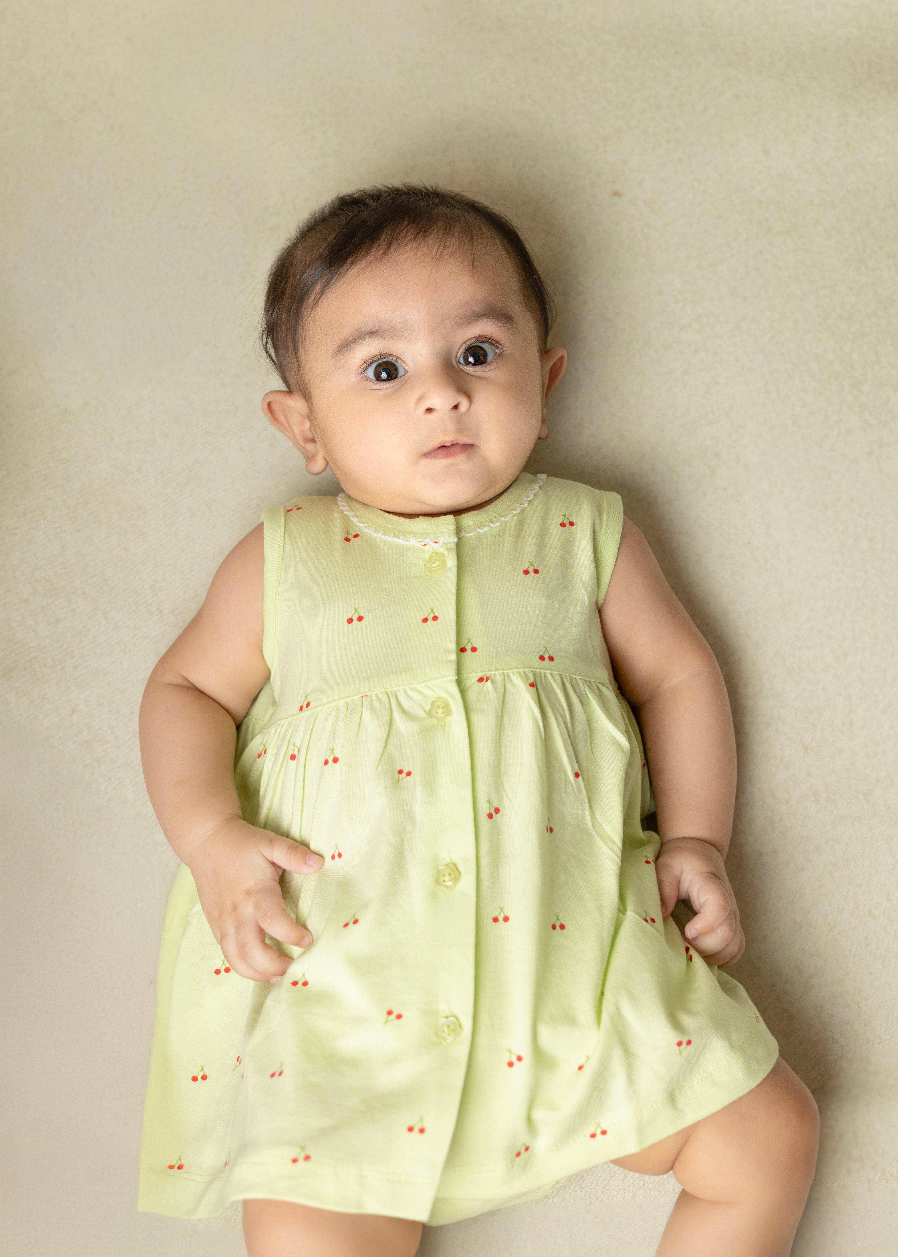 A baby wearing a light green sleeveless cotton frock with cherry blossom prints and a matching bloomer.