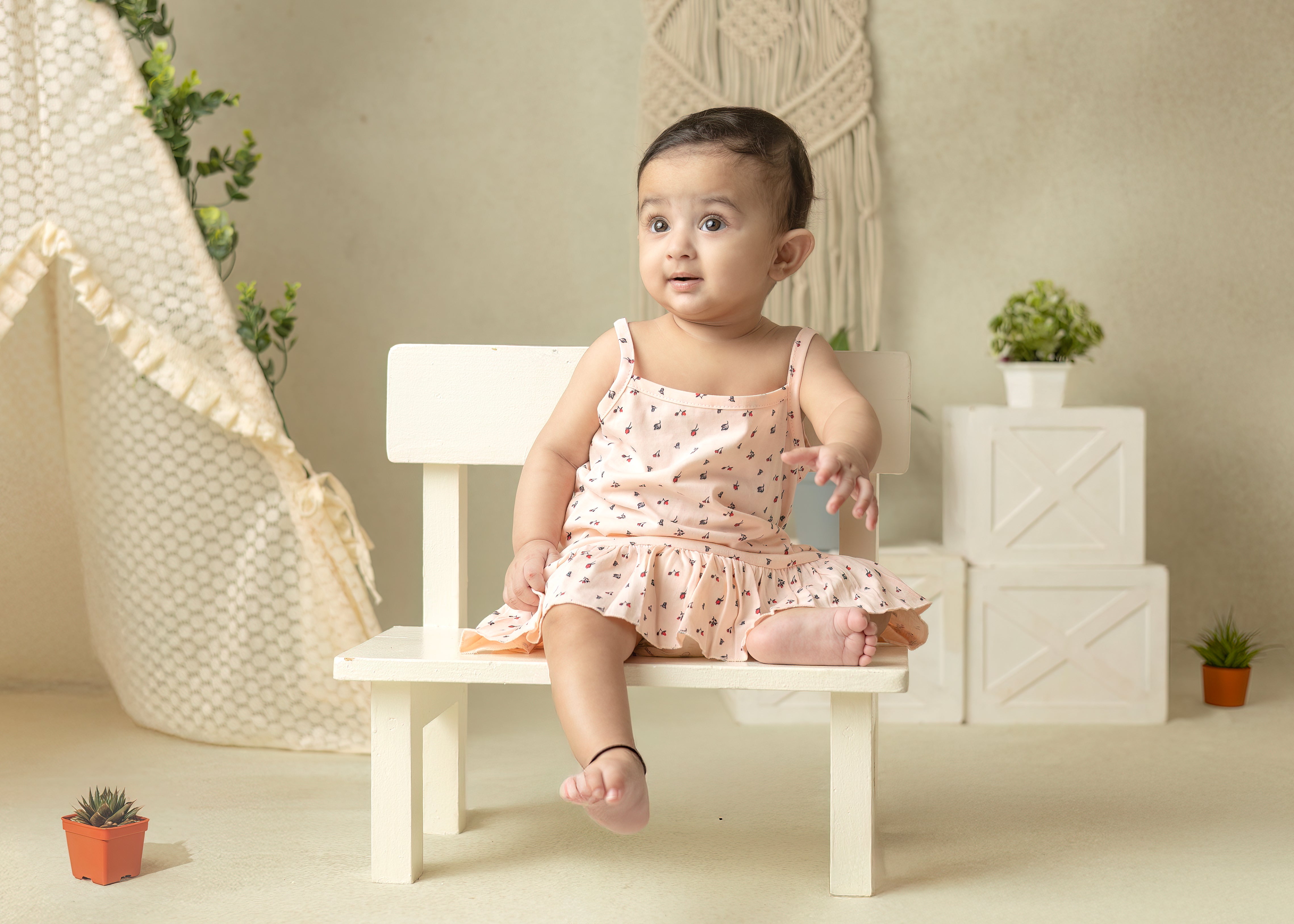 A baby wearing a floral frills frock in peach blush color, sitting on a small chair with a decorative room setup.