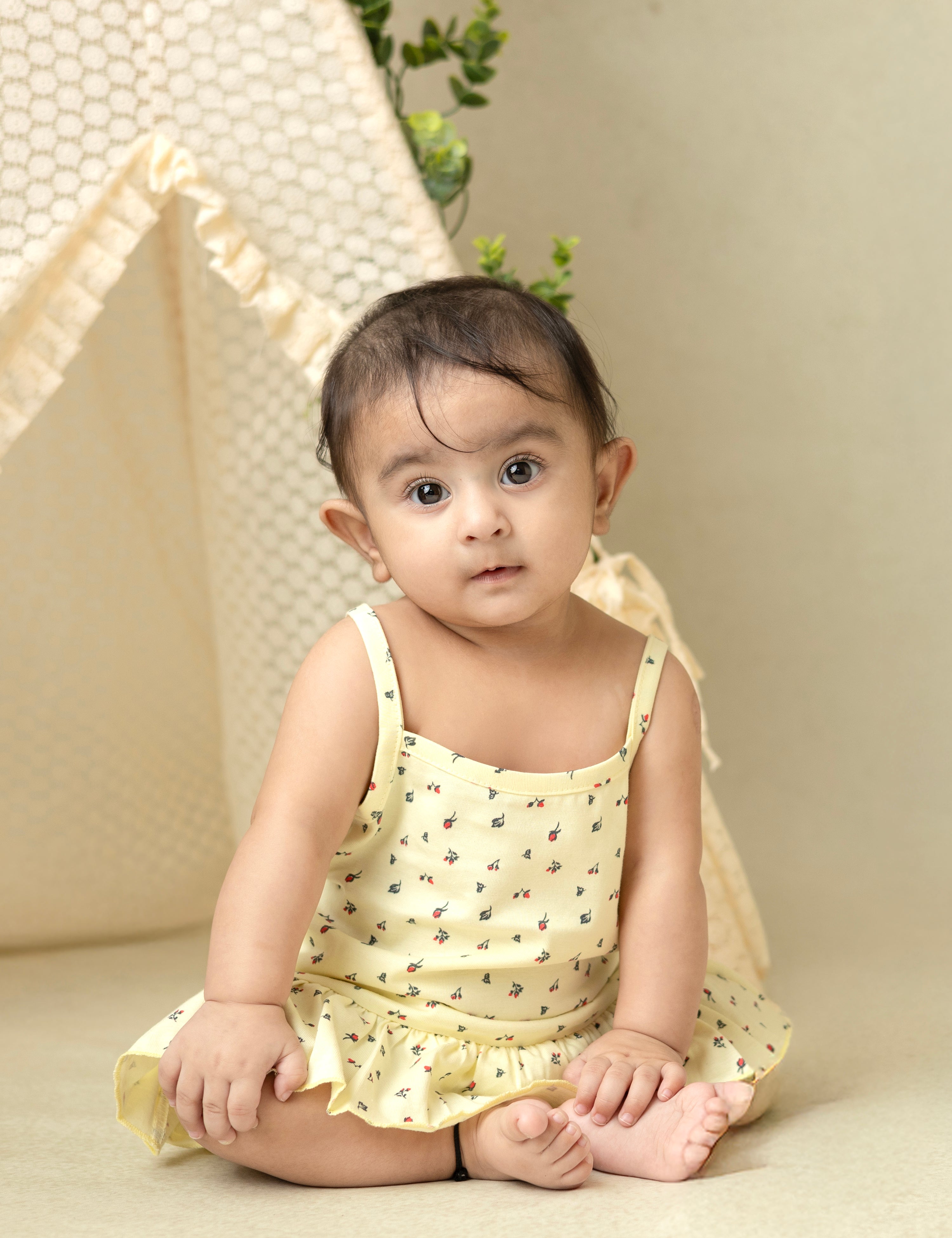 A baby wearing a beige cotton frock with a floral design and frill detailing at the hem, sitting against a beige background.
