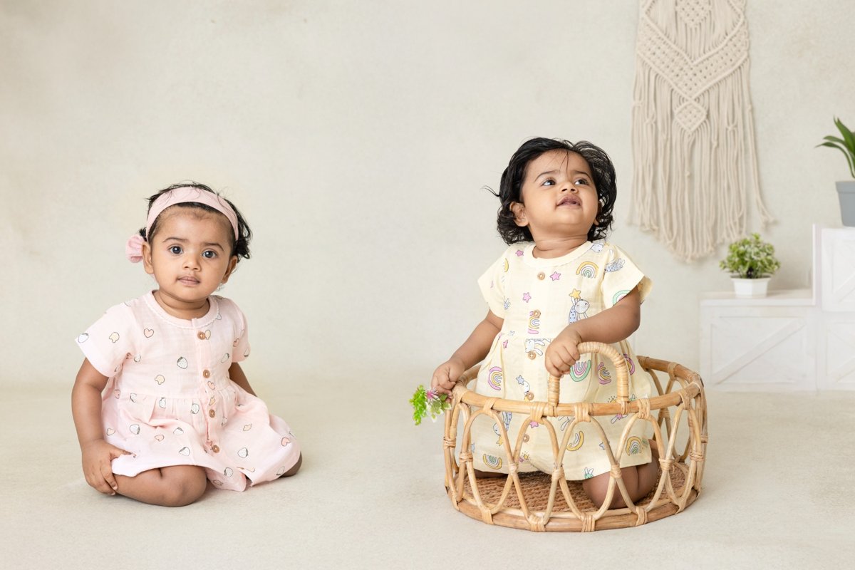 Two young children in light-colored outfits sitting on a white floor with a minimal background.