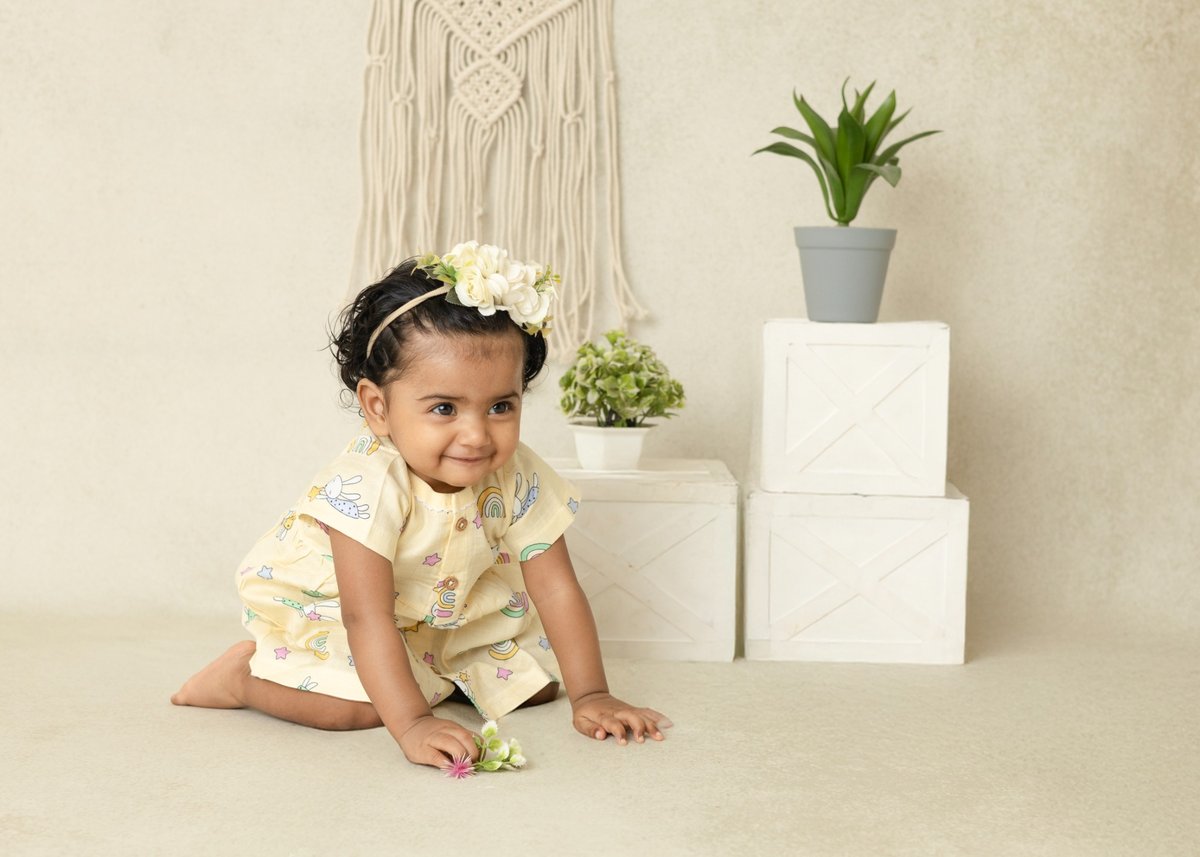 Baby in a floral dress sitting on the floor with plants and decorative items in the background