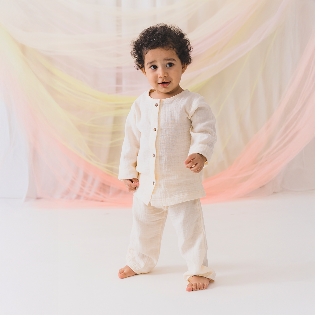 Child wearing a white outfit standing in front of a soft, multicolored curtain.