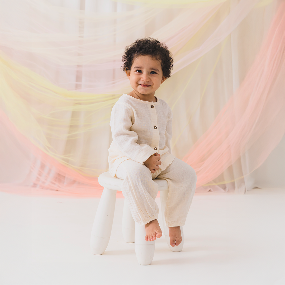 Child sitting on a stool wearing a white outfit against a soft, multicolored curtain background